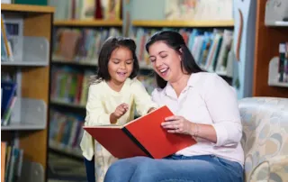 A woman and a young girl happily read a large book together in a library, surrounded by bookshelves.