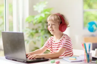 A young child wearing red headphones uses a laptop at a desk with school supplies, in a bright room.