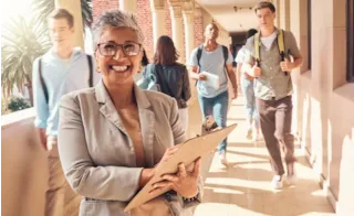 Smiling woman with clipboard stands in school hallway as students walk past her in the background.