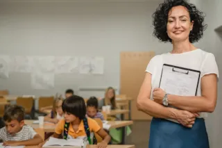 Smiling teacher holding clipboard stands in classroom while students work at desks in the background.