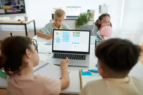 Four children sit around a table, working on laptops and notebooks, focused on a coding project.