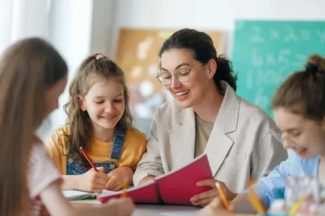 A teacher helps three young students with their schoolwork at a table in a classroom, all smiling and engaged.