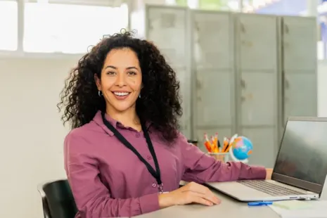 Smiling woman with curly hair sits at a desk with a laptop in a classroom, lockers and supplies in the background.