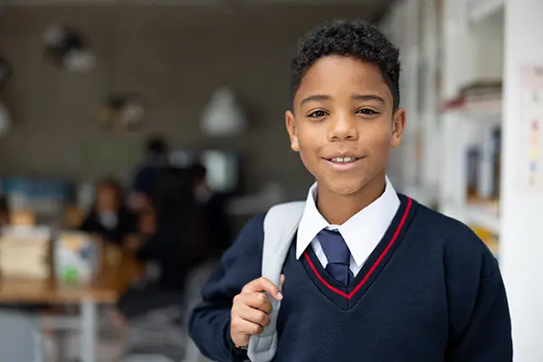 Smiling schoolboy in a uniform and backpack standing in a classroom, looking at the camera.