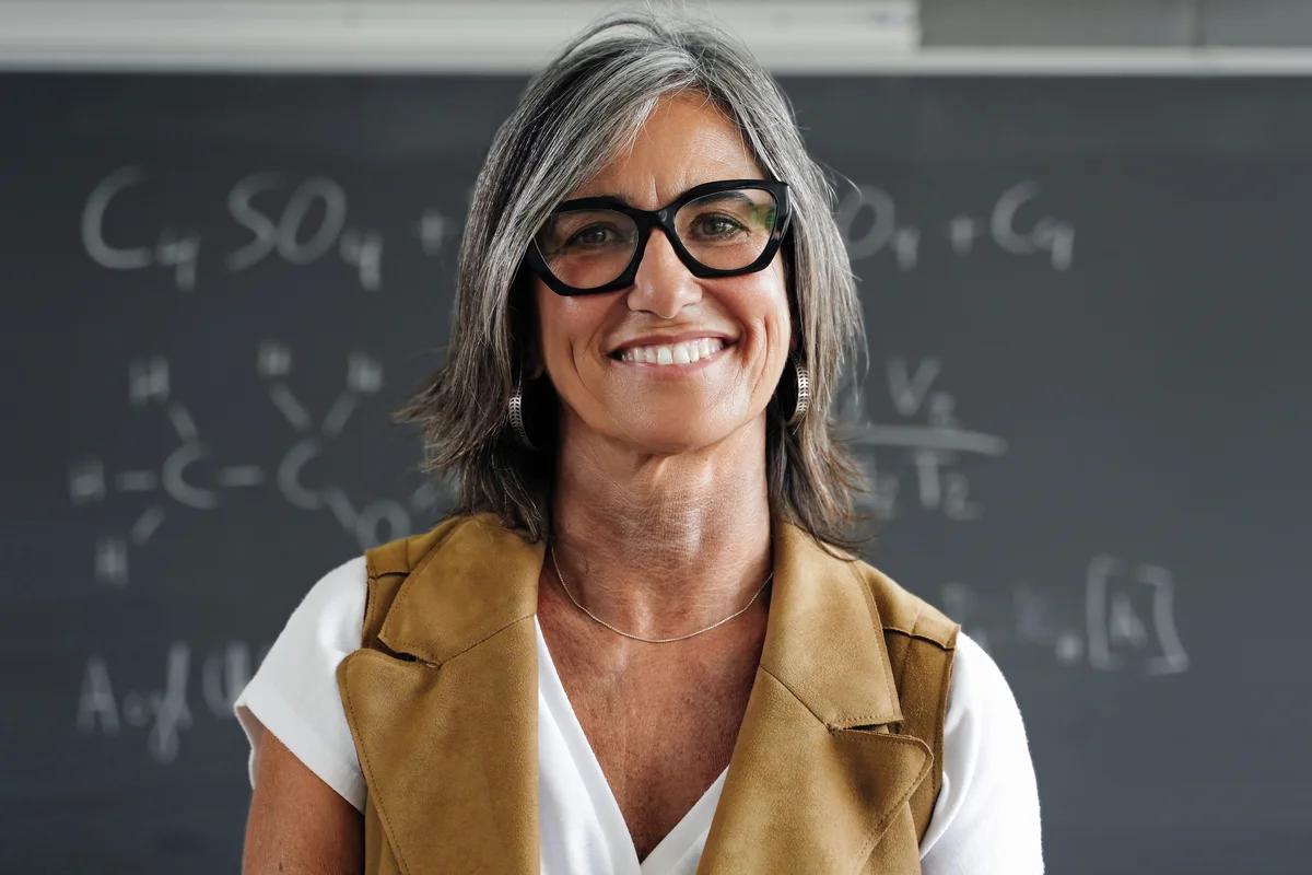 Smiling woman with glasses stands in front of a chalkboard with chemistry formulas written on it.