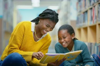 Two girls sitting on a library floor smiling and reading a book together, surrounded by bookshelves.