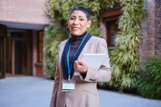 Smiling person in a tan blazer holds a tablet and wears a lanyard, standing outside near green plants and brick walls.
