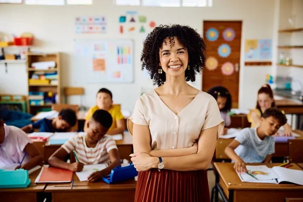 Smiling teacher stands in a classroom with students working at their desks in the background.