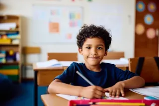 Smiling child sits at a classroom desk, coloring with markers and paper, with a whiteboard in the background.