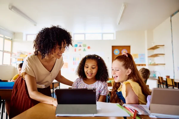A teacher helps two young girls who are smiling and looking at a tablet in a bright classroom.