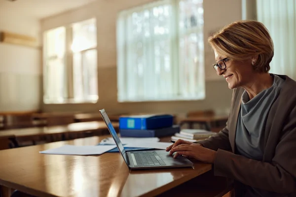 Smiling woman using a laptop at a desk in a sunlit classroom with papers and binders nearby.