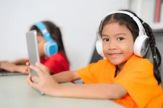 Smiling girl in orange shirt using tablet with headphones; another child with headphones in the background.
