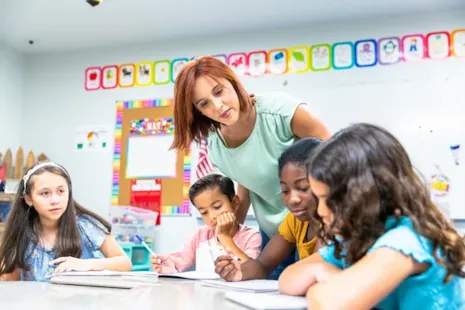 A teacher helps a group of four young students working on assignments at a table in a colorful classroom.