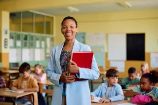 Smiling teacher holding a red folder stands in front of students working at desks in a classroom.