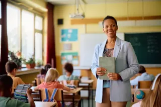 Smiling teacher holding a book stands in front of students sitting at desks in a bright classroom.