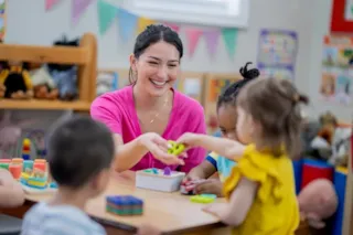 A teacher smiles as she plays with three young children at a table in a colorful classroom.