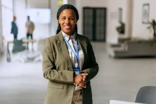 Smiling woman in a business suit with a lanyard, standing in a modern office environment.