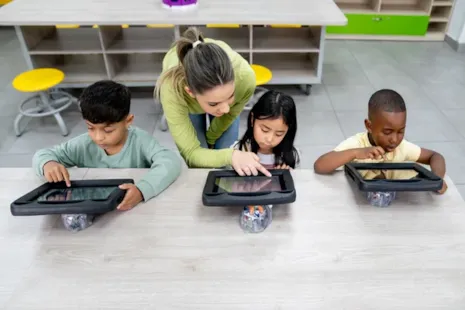 A teacher helps three young children using tablets at a classroom table with chairs and desks in the background.