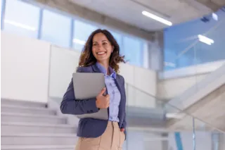 Smiling woman in business attire holds a laptop while standing near modern office stairs.