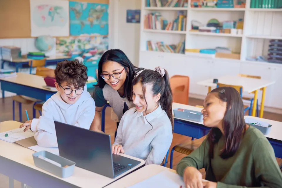 A teacher and three students sit at a desk, smiling and looking at a laptop in a classroom.