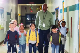 A teacher walks down a school hallway with a diverse group of smiling young students wearing backpacks.