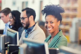 Smiling woman looks at the camera while working at a computer in a row with other people in an office.