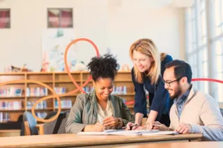 Three adults sit and collaborate at a library table, smiling and working together with books and papers.