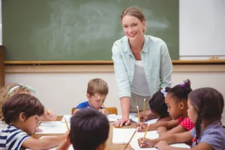 Smiling teacher stands by a desk while young children write in notebooks in a classroom with a chalkboard.