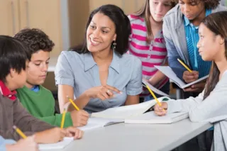 A teacher sits at a table with five students, engaging in a discussion. Students hold notebooks and pencils, leaning in attentively.