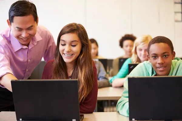 Teacher helps a smiling student with her laptop while other students work in a classroom setting.