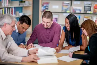 Five adults sit together at a table in a library, reading and writing, appearing to work on a group project.