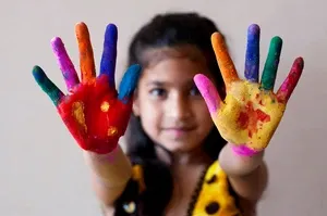 A girl smiles, holding up her hands covered in bright, multicolored paint, with a neutral background.