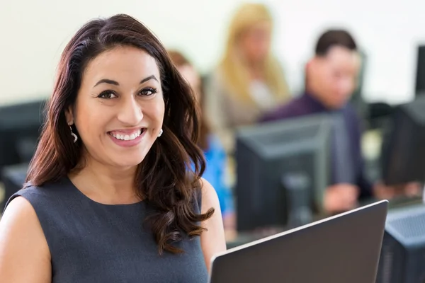Smiling woman in business attire at a computer in an office, with colleagues working in the background.