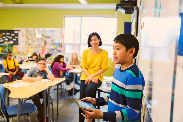 A boy presents with a tablet in front of a classroom while students and a teacher watch and listen.
