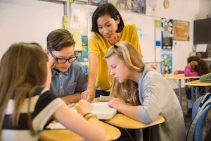A teacher helps three students with their work at a round classroom table. Other kids work in the background.