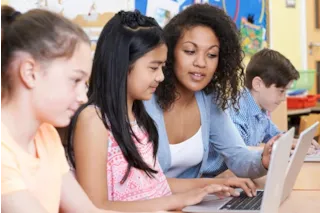 Teacher assisting students working on laptops in a classroom. The room has colorful bulletin boards in the background.