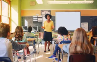 Teacher stands at the front of a classroom, speaking to students sitting at desks facing her.