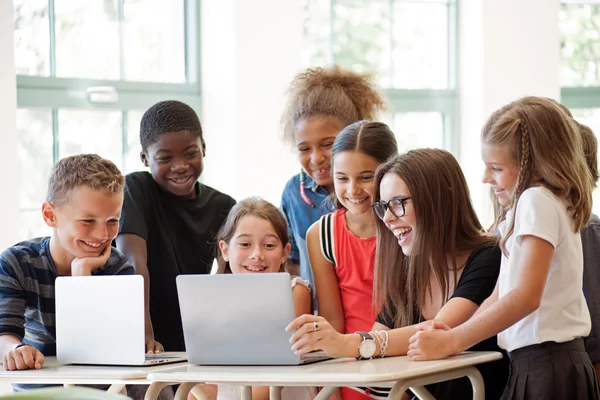 A group of smiling children and a teacher gather around a table, looking at laptops in a bright classroom.