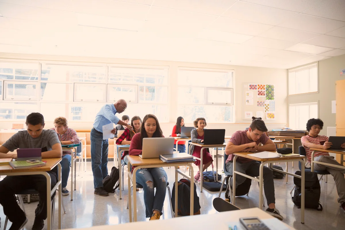 High school students sitting at desks using laptops and tablets in a bright classroom while a teacher assists a student.