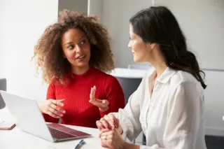 Two women sit at a table with a laptop, having a conversation in a bright, modern office setting.