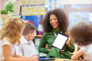 Smiling teacher shows a blank tablet to three young students in a classroom setting.