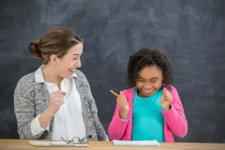 A teacher and student celebrate a success together at a desk in front of a chalkboard.
