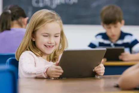 Smiling young girl sits at a desk using a tablet in a classroom, with other students working in the background.