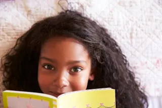 Young girl with curly hair smiles while reading a yellow book, partially hiding her face, on a patterned bedspread.