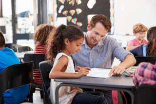 A teacher reads with a student seated at a desk.