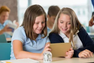 Two girls in school uniforms smiling while looking at a tablet in a classroom setting, with other students in the background.