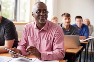 A man in a pink shirt sits at a desk in a classroom, looking to his side.