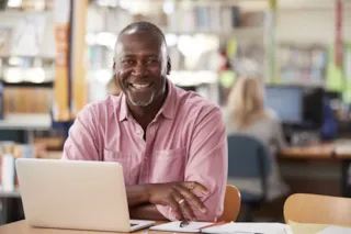 Smiling man in a pink shirt sits at a desk with a laptop in a library or study area, with bookshelves in the background.
