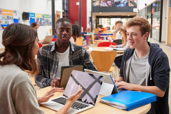 Three students sit at a table with laptops and notebooks, talking and studying in a bright, modern school common area.