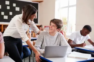 A teacher leans over to help a smiling student with a laptop in a classroom while other students work.